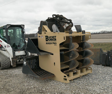 Auger PRO rack loaded with auger drive, three auger bits, buckets, and pallet forks beside a skid steer.